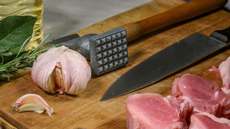 Head of garlic and clove with pork on cutting board with chef knife and meat tenderizer.