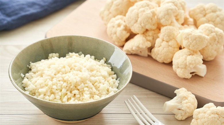 A bowl of cauliflower rice next to raw cauliflower florets.