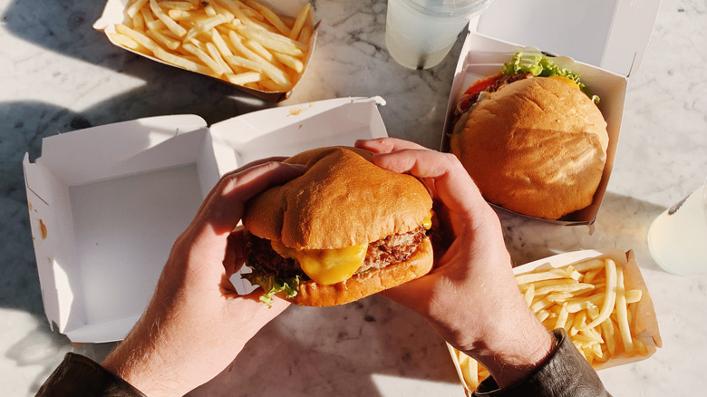 Hands holding burger with another burger and fries in the background