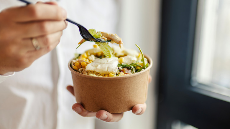 A closeup of a person holding a forkful of chicken and corn salad in one hand, and the disposable salad container in another