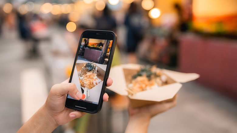 A person holding up a takeout box with one hand and taking a photo of it with a phone on the other