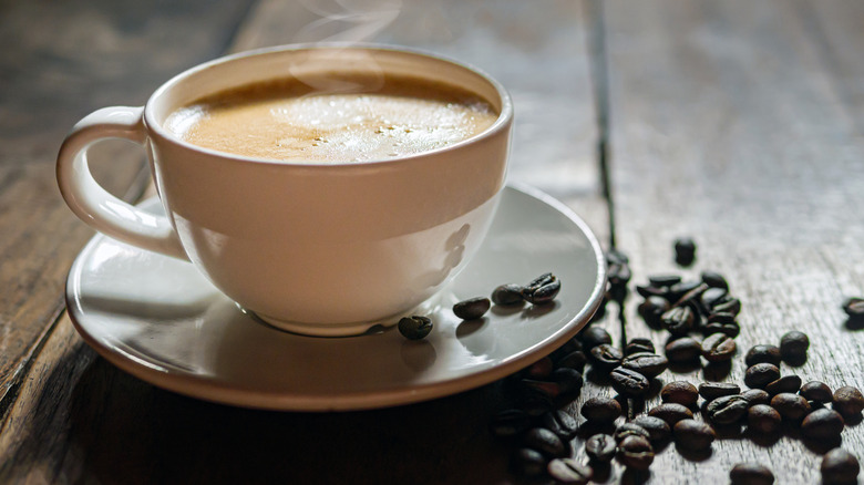 A cup of coffee on a wooden table surrounded by coffee beans.