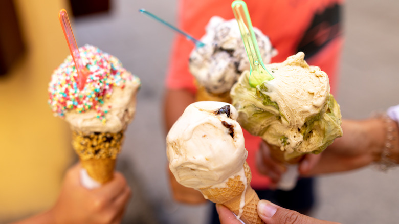 Four hands holding up ice cream cones, three of which have plastic spoons in them.