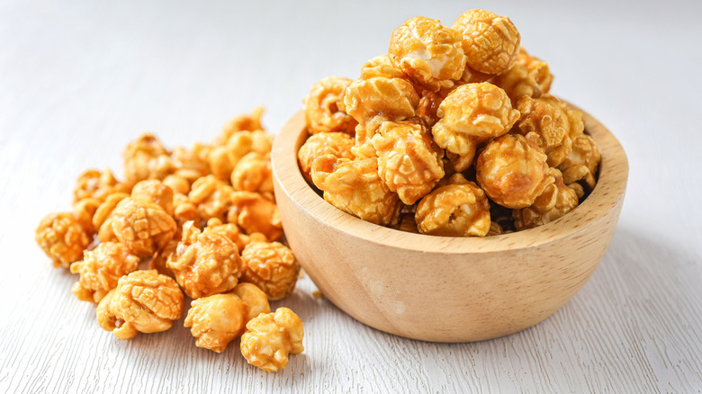 Caramel popcorn in a wooden bowl on a white table, a pile of caramel corn next to the bowl.