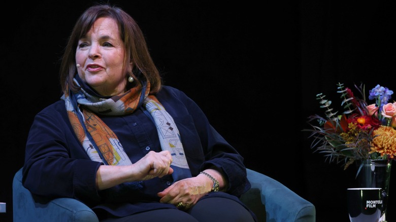 A seated Ina Garten speaks onstage at a live event with flowers nearby.