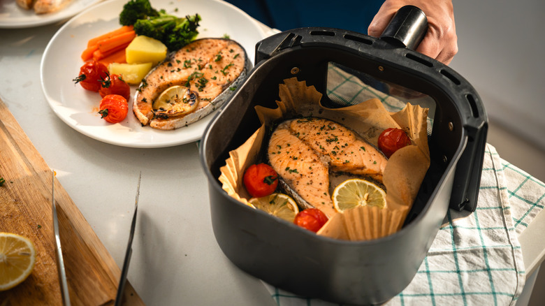 Hand holding an air fryer basket filled with fish, lemon, and tomatoes.