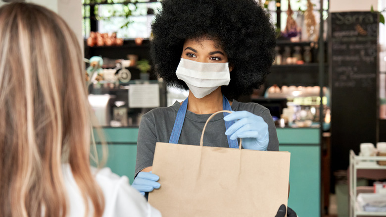 A woman wearing a mask and handing a paper bag to someone else