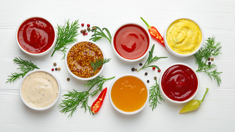 Seven bowls of various sauces on a table, surrounded by various herbs and peppers.