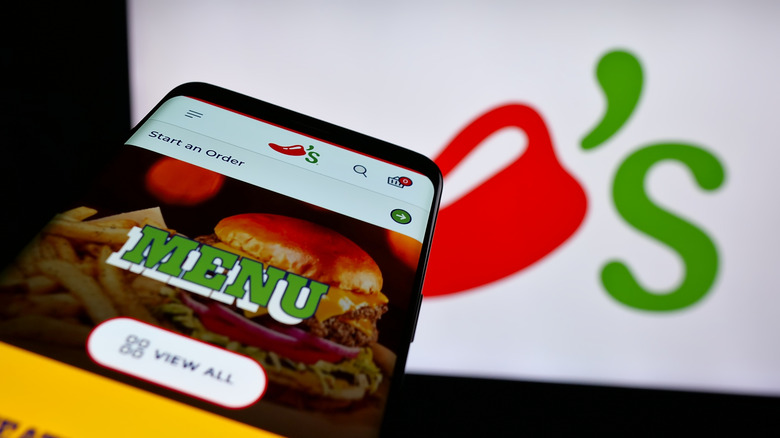 A mobile phone showing the menu header of Chili's website with the restaurant's logo on a screen in the background
