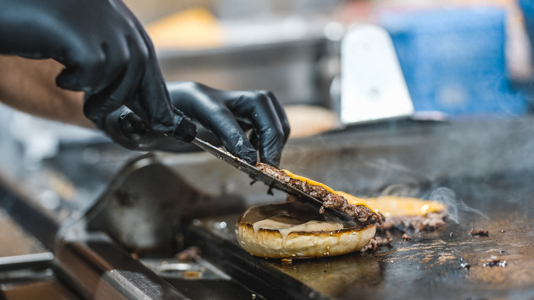 Closeup of hands with black gloves laying a grilled burger patty on top of a bun on a griddle