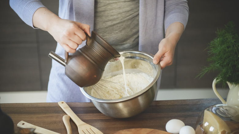Pouring milk from ceramic jug into metal bowl with flour