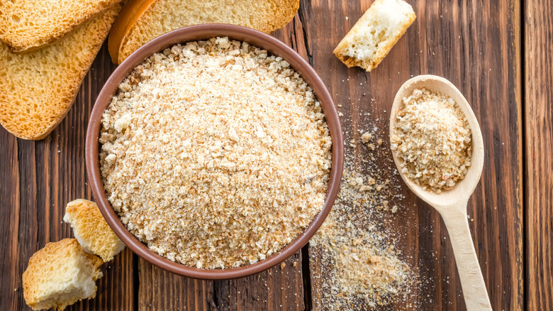 A bowl of breadcrumbs with spoon and slices of bread