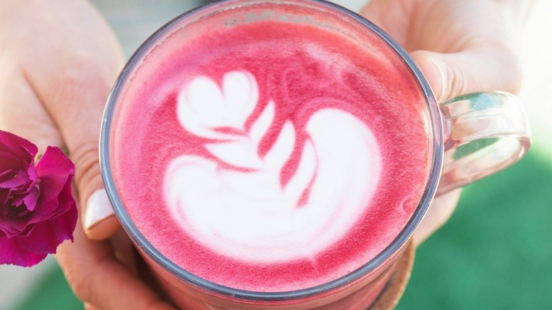 Hands holding a pink latte in a cup, along with a flower