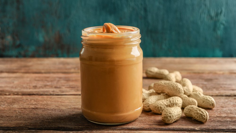 glass jar of peanut butter with whole peanuts next to it on a wooden table