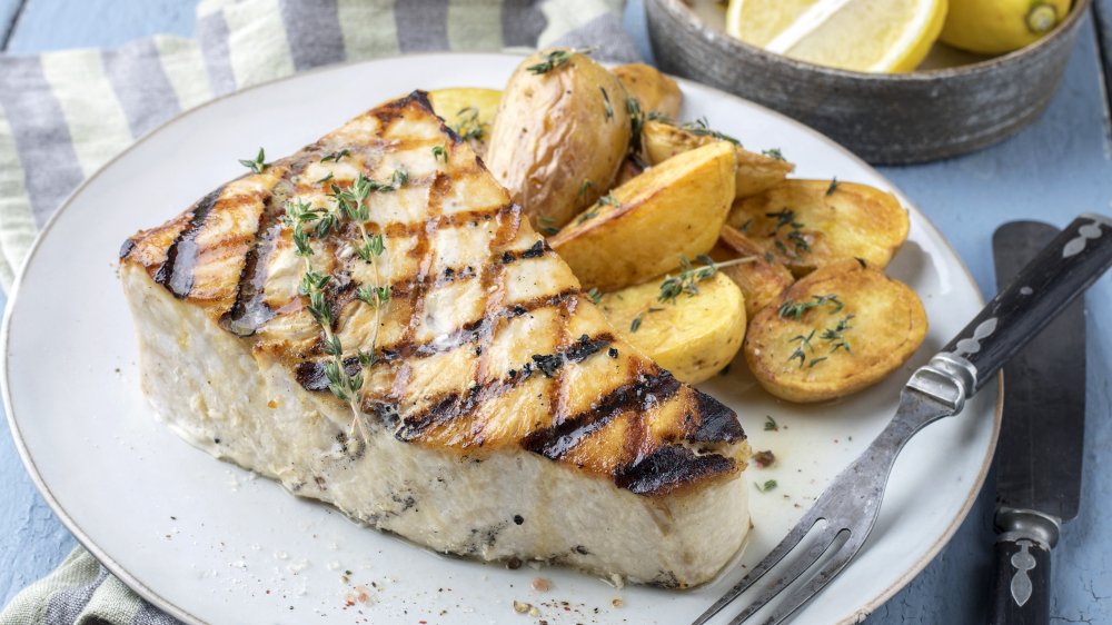 Thick piece of grilled swordfish on a white plate next to potatoes, twigs of thyme on top of both, a seafood fork is on edge of plate