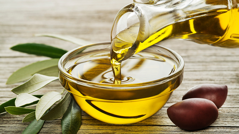 Bottle of Olive oil pouring in a glass bowl with olives and branch