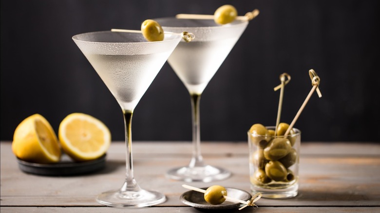 two martinis in cocktail glasses with olives on a table with a jar of olives and a cut lemon