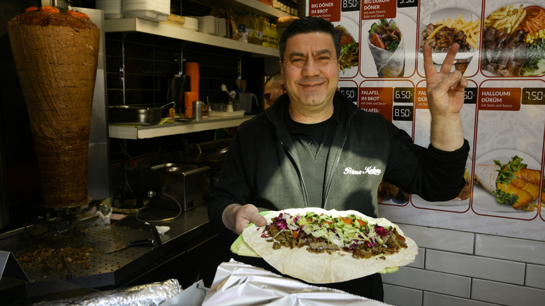 street food stall owner giving peace sign, holding kebab mid-assembly