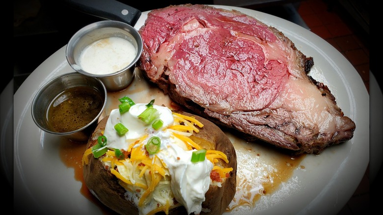 A slice of prime rib with au jus and horseradish sauce, served with a baked potato topped with sour cream, cheddar cheese, green onions, and bacon