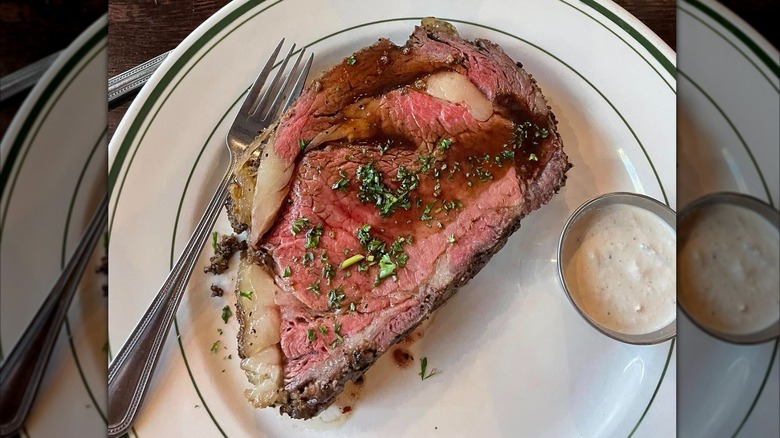 A slice of prime rib topped with au jus and herbs, served with horseradish sauce and a loaded baked potato