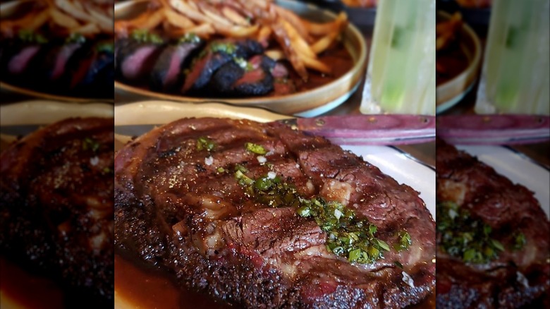 A slice of prime rib topped with herbs and sauce, and a dish with fries and a sliced steak in the background