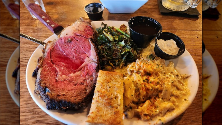 A slice of prime rib served with cooked greens, potatoes, and au jus