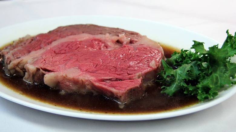 A slice of prime rib in au jus served with a side of greens on a white plate