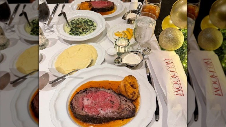 A dinner table with two plates of prime rib, a bowl of creamed spinach, a bowl of mashed potatoes, and glasses of water on white tablecloth