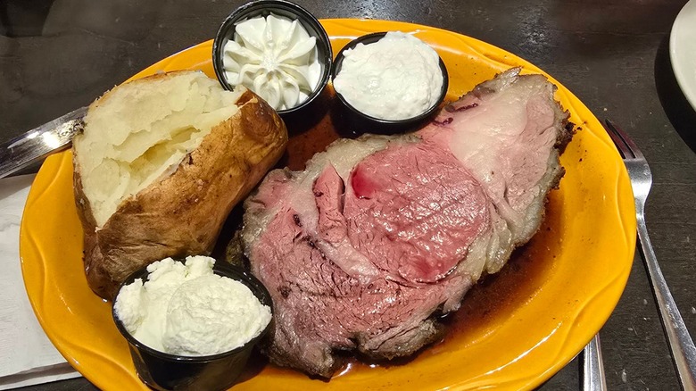 A full yellow plate with a piece of prime rib, a baked potato, and sides of butter, sour cream, and horseradish sauce