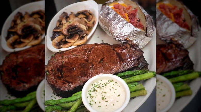 A piece of prime rib on a plate with a loaded baked potato, sautéed mushrooms, asparagus, and a creamy sauce
