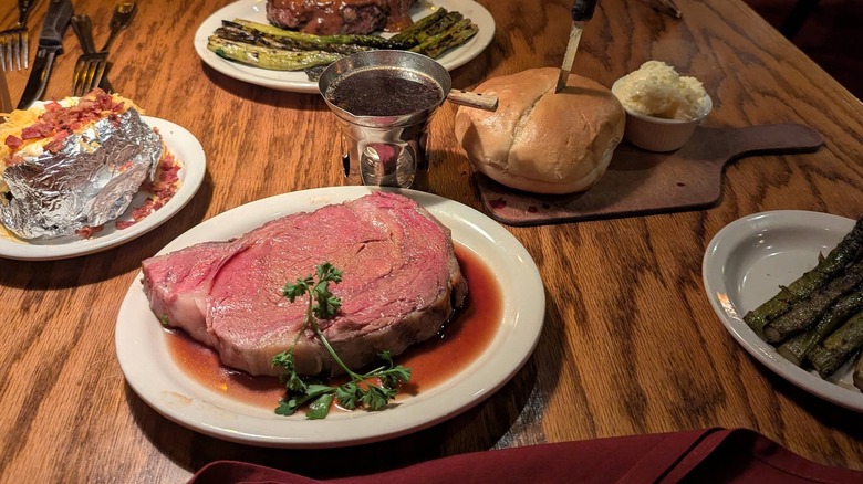 A large piece of prime rib on a white plate in au just surrounded by other plates of sides, including a loaded potato, asparagus, and fresh bread and butter