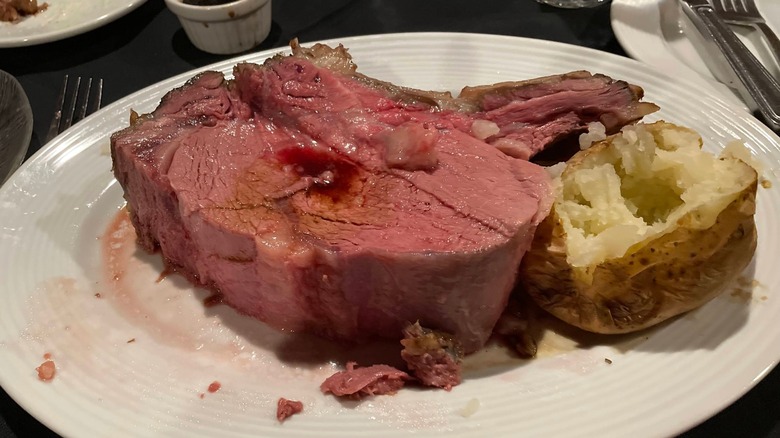A thick slice of prime rib next to a baked potato on a white plate
