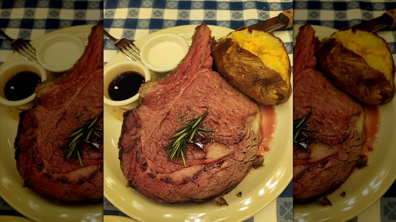 A large piece of prime rib garnished with a sprig of rosemary with a baked potato and sides of au jus and horseradish sauce