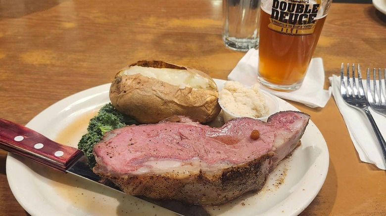 A slice of prime rib with a baked potato and horseradish sauce with a beer next to the plate