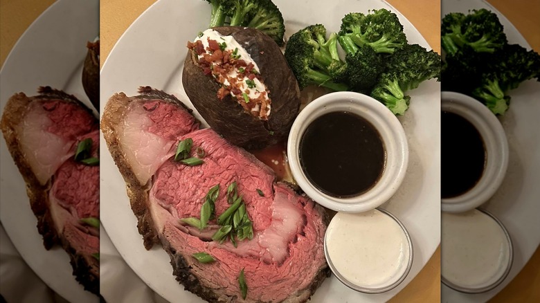 A slice of prime rib with au jus, horseradish sauce, a loaded baked potato, and broccoli