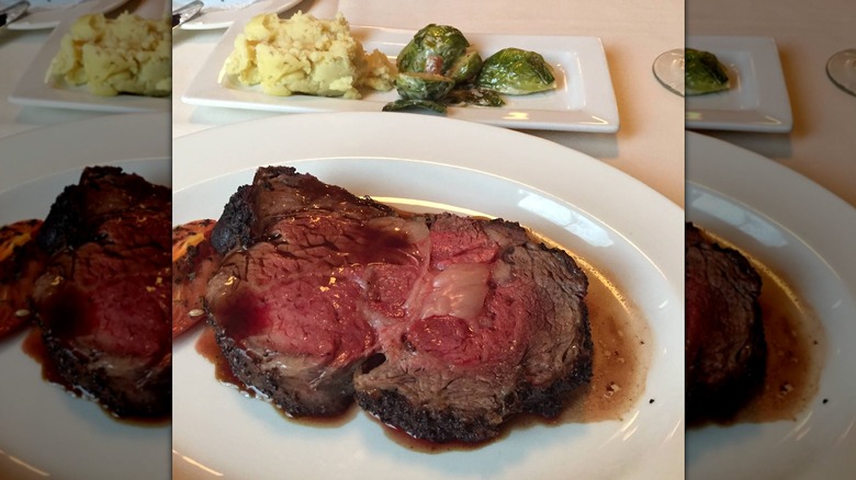 A slice of prime rib in its own au jus on one plate and mashed potatoes and Brussels sprouts on another plate