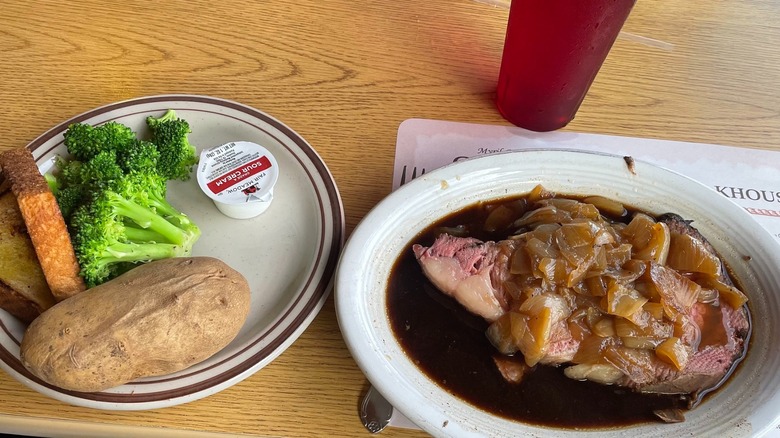 A slice of prime rib in its own au jus and sautéed onions on one plate and a baked potato and broccoli on another plate