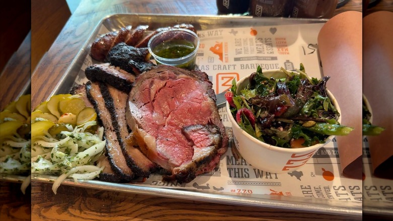 Thin sliced prime rib served on a BBQ platter with sausage, pickled veggies, greens, and a chimichurri sauce