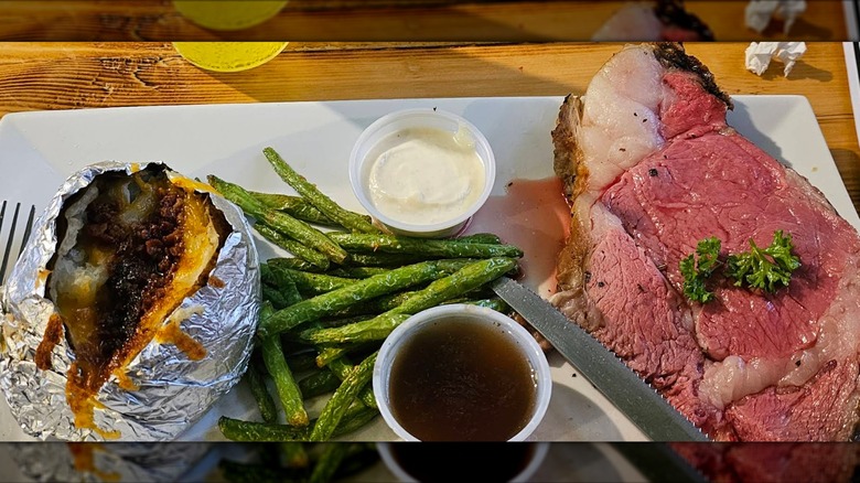 A pink slice of prime rib served with au jus, horseradish sauce, a baked potato, and green beans