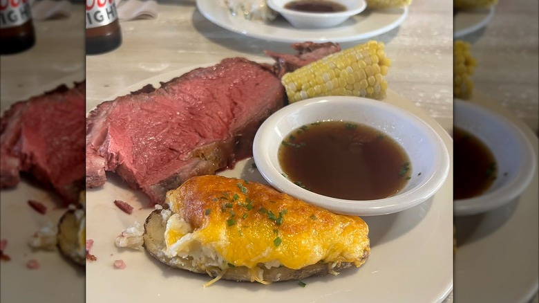 A thick slice of prime rib served with au jus, a baked potato, and half a corn on the cob