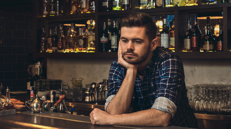 Pensive bartender leaning on counter with many liquor bottles behind him