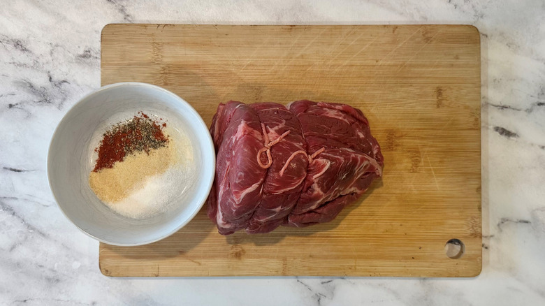string-wrapped raw chuck roast on a wooden board next to a bowl of seasonings