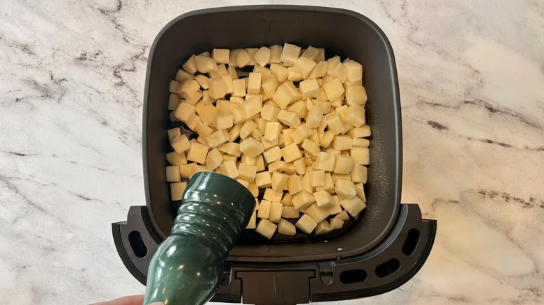 hand holding a green pepper grinder over an air fryer full of potato cubes
