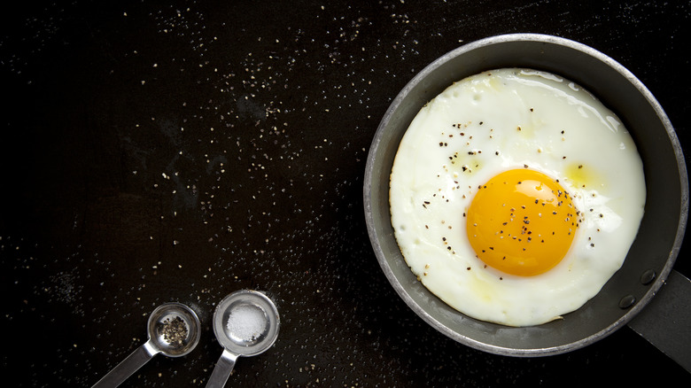 A fried egg on a skillet, next to spoons with salt and pepper, over a dark background