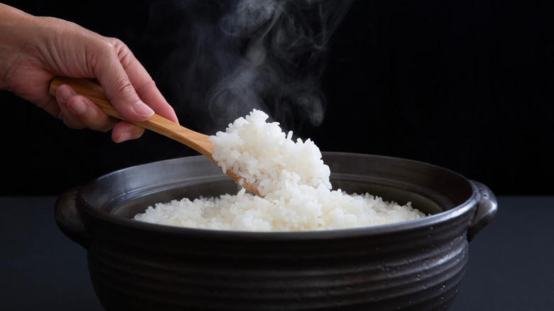 A person spoons cooked white rice from a black bowl with a wooden spoon.