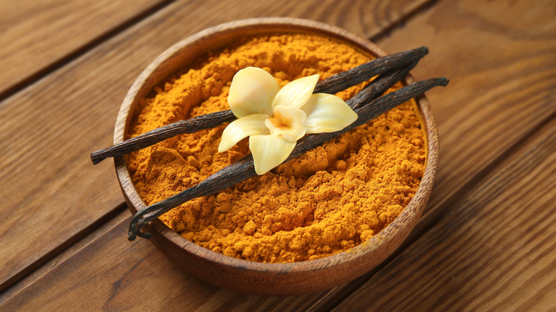 Bowl of turmeric powder with vanilla pods on top and a flower