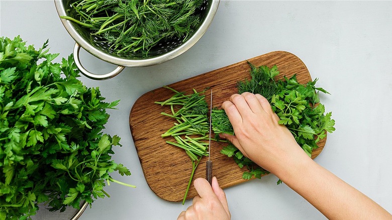 Hands chopping herbs in a wooder board, next to strainers with more herbs 