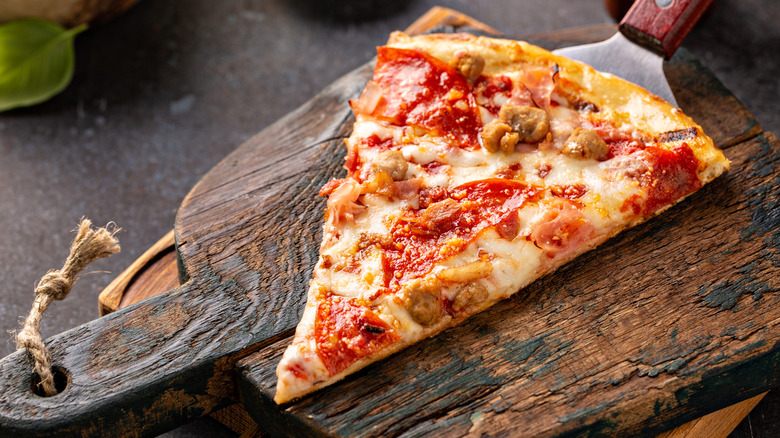 A slice of eat lover's pizza on wooden cutting board