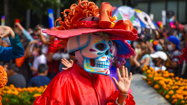 A woman in a red dress and hat with a colorfully painted skull mask marches in a Día de los Muertes parade.