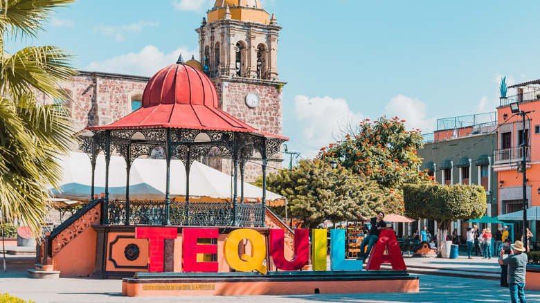 Tourists take pictures of the colorful letters that spell out "Tequila" with historic stone buildings in the background.